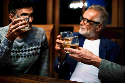 Two men clink whiskey glasses at a warm, dimly lit table, smiling in conversation, with one man blurred in the foreground